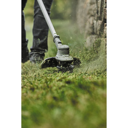 Person uses a DEWALT string trimmer to cut grass near a stone wall outdoors.