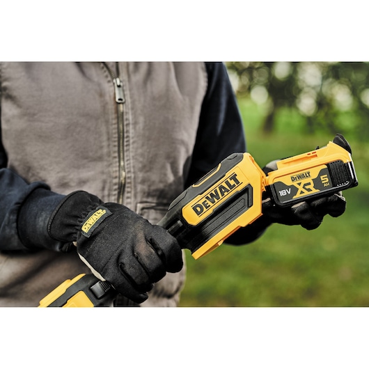 Close-up of gloved hands holding a DEWALT 18V XR cordless tool outdoors, showing battery and branding.