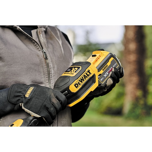 Person holding a DEWALT cordless tool, wearing gloves and a zip jacket, outdoors in a garden setting.