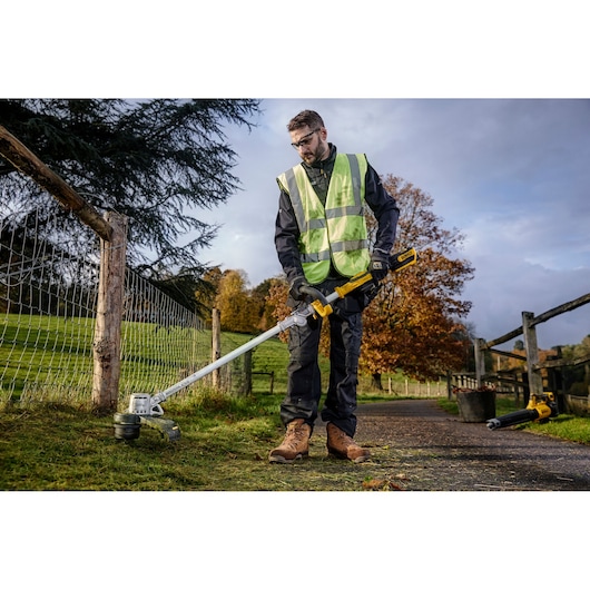 Person using a DEWALT cordless string trimmer outdoors near a fence in a grassy area.