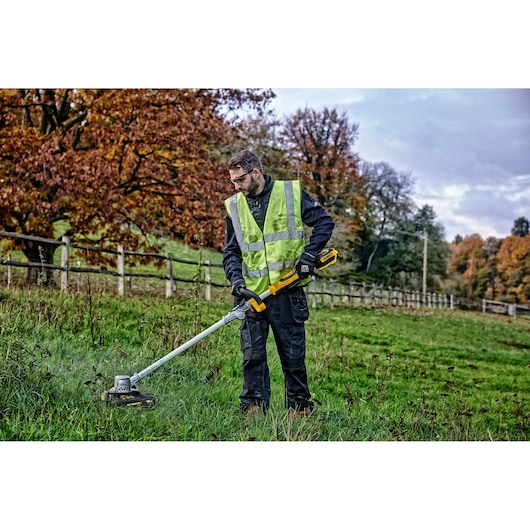 Person using a DEWALT cordless grass trimmer outdoors, wearing a safety vest and gloves in a grassy area.