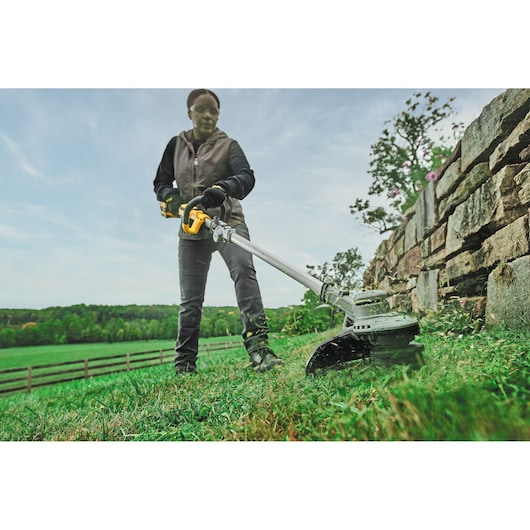 Person using a DEWALT cordless string trimmer to cut grass near a stone wall outdoors.