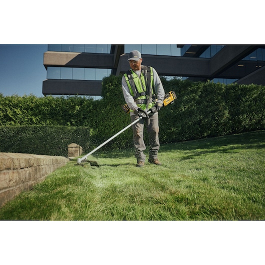 Person trims grass with a DEWALT DCMST901N cordless string trimmer near a brick edge in a landscaped area.