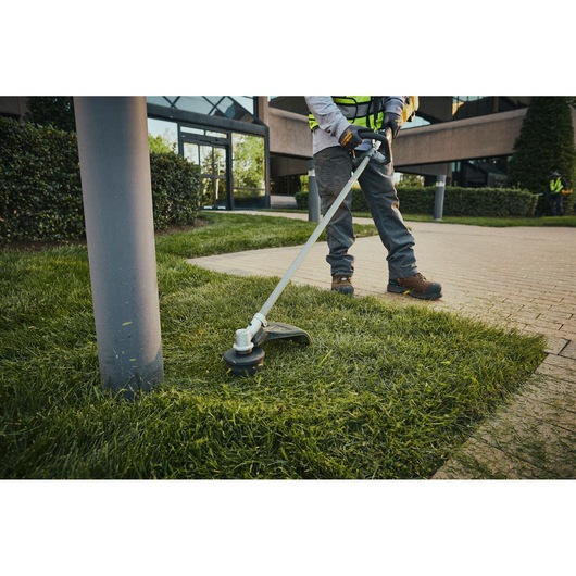 Person using DEWALT cordless grass trimmer near sidewalk and lamp post in landscaped outdoor area.