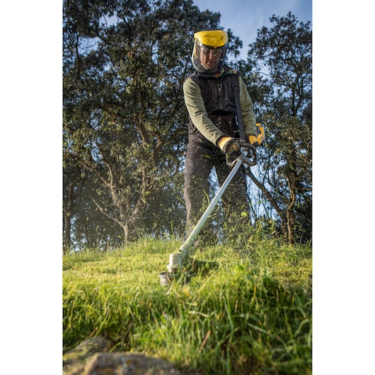 Person uses DEWALT grass trimmer outdoors, wearing safety gear on grassy area with trees in background.