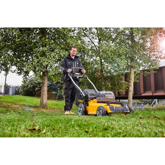 Person using a DEWALT DCMW564N lawn mower on grass outdoors, surrounded by trees and buildings.