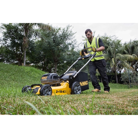 Man uses a DEWALT DCMW564N lawn mower to cut grass outdoors, wearing a high-visibility vest and gloves.