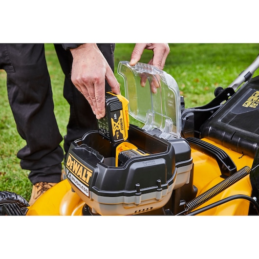 Person inserts DEWALT battery into yellow lawn mower compartment outdoors on grass.