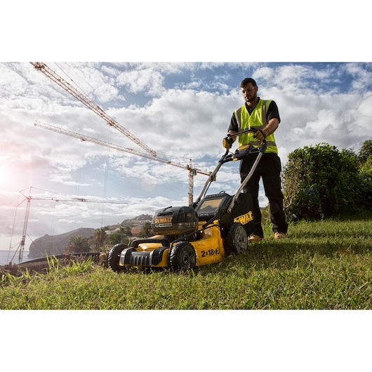 Person uses a DEWALT lawn mower on grass, with construction cranes and cloudy sky in the background.