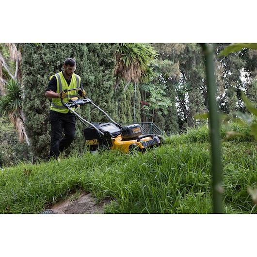 Person uses a DEWALT lawn mower on a grassy slope in a garden surrounded by trees.