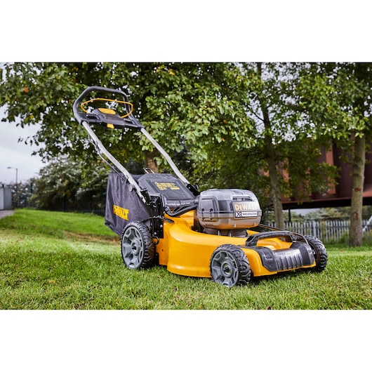 DEWALT cordless lawn mower on grass outdoors, yellow and black, with trees in the background.