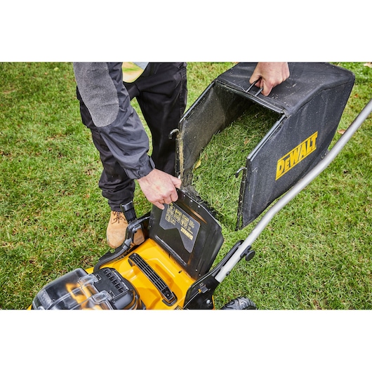 Person removes grass catcher from DEWALT lawn mower on green lawn.