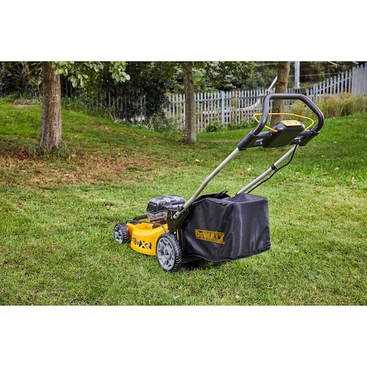 DEWALT cordless lawn mower on grass with trees and a fence in the background.