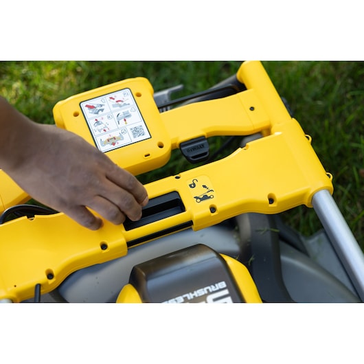 Close-up view of a hand adjusting a yellow DEWALT lawn mower handle with visible instructional stickers showing usage steps, placed on grass.