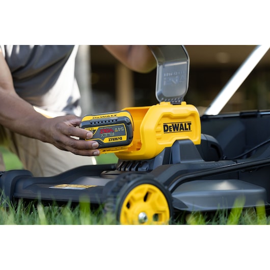 A person inserts a DEWALT FLEXVOLT battery into a DEWALT cordless lawn mower, with the mower positioned on grass.