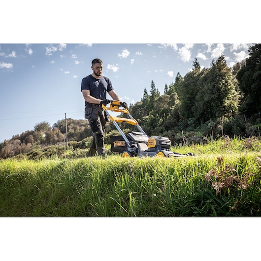 Person mowing grass outdoors with a DEWALT lawnmower on a sunny day, trees and blue sky in the background.