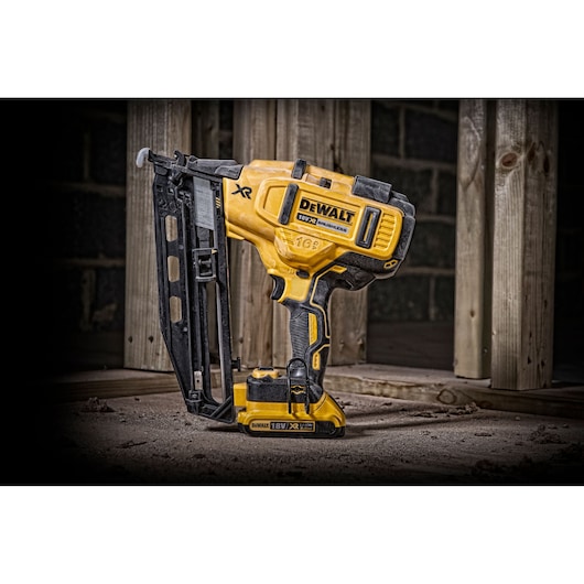 DEWALT 18V cordless nail gun stands on a dusty floor near wooden beams at a construction site.