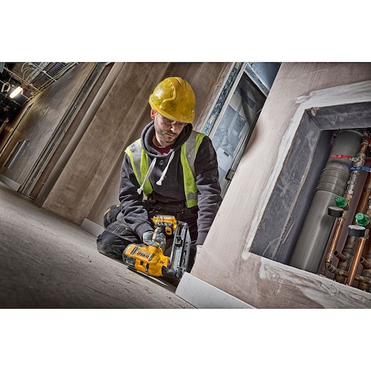 Worker kneeling indoors using a DEWALT DCN660D2 nailer on baseboards. Wearing a yellow hard hat and safety vest.