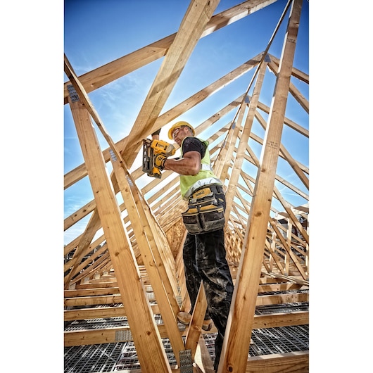 Worker uses a DEWALT nailer for wood construction on a roof, wearing safety gear and tool belt.