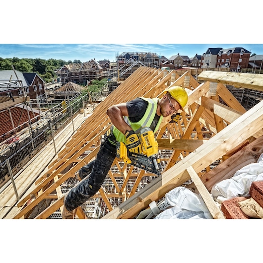 Worker uses a DEWALT nailer on wooden roof frame at a construction site with houses in the background.