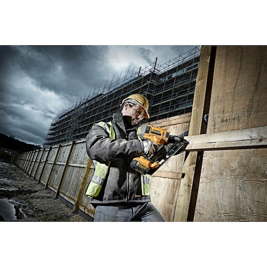 Person uses DEWALT nailer DCN692P2 near wooden fence at a construction site wearing safety gear.