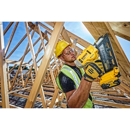 Worker uses DEWALT DCN692P2 nailer on wood frame construction, wearing safety gear under blue sky.