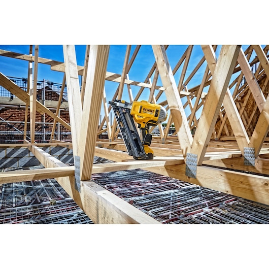 DEWALT nailer tool placed on wooden beams at a construction site with trusses and blue sky in the background.