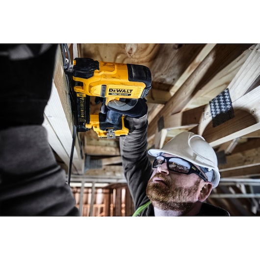 Person using a DEWALT DCN701D2 tool on ceiling framework at a construction site, face blurred for privacy.