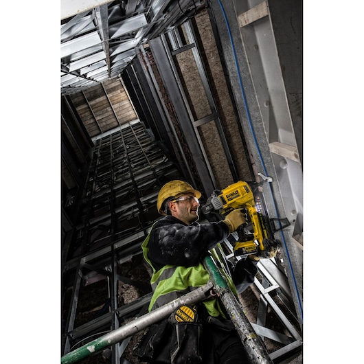 Worker using DEWALT DCN890P2 nailer on metal framework, wearing safety gear in construction site.