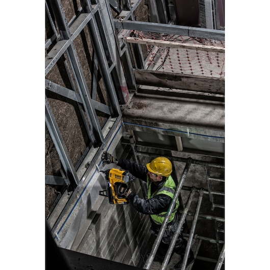Worker uses DEWALT DCN890P2 nailer on wall frame at a construction site, wearing safety gear.