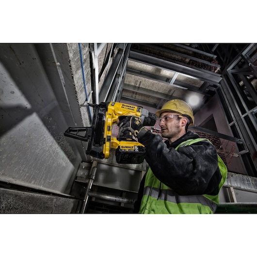 Worker uses DEWALT DCN890P2 nailer on metal frame in industrial setting, wearing safety gear and helmet.