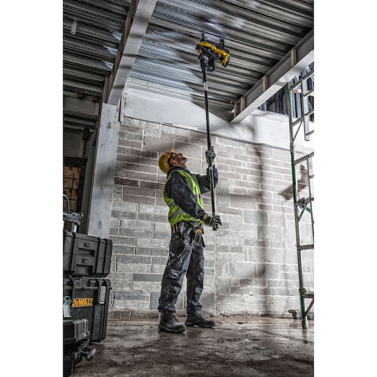 Worker uses DEWALT DCN890P2 tool overhead at construction site. Safety gear and tool cases visible.
