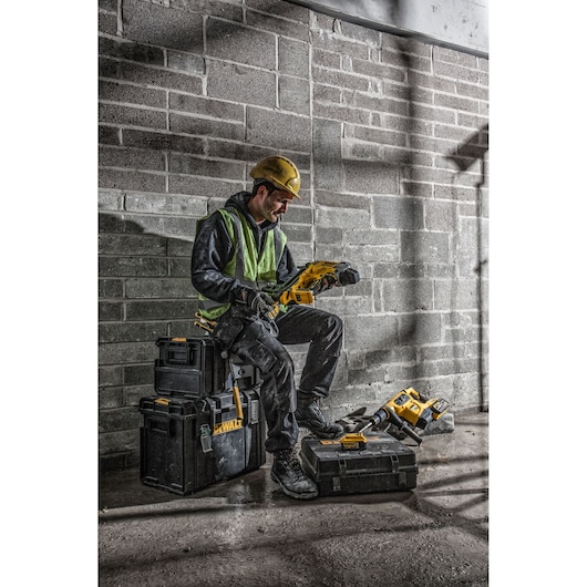 Worker in safety gear using a DEWALT tool, sitting on DEWALT boxes in a construction setting with brick wall.