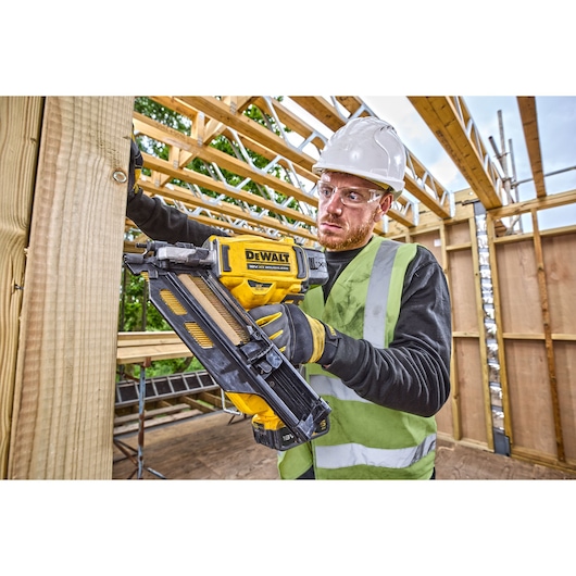 Person using a DEWALT DCN930N nailer in construction, wearing safety gear and working with wooden beams.