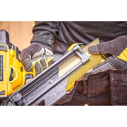 Gloved hands loading nails into a yellow DEWALT 18V XR brushless nail gun at a construction site.