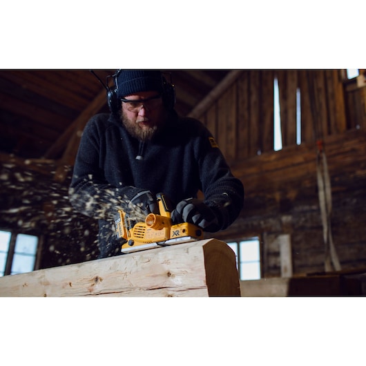 A person using a DEWALT DCP580P2 cordless planer to smooth a piece of wood inside a workshop, with wood shavings flying. The person's face is obscured and they are wearing protective headphones and gloves.