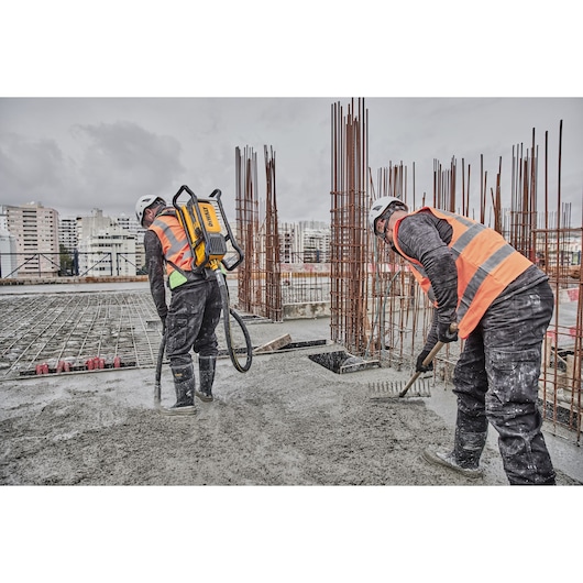 Two workers in safety gear use a DEWALT tool on a construction site with concrete and steel rebar.