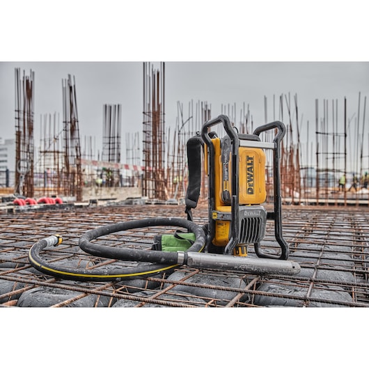 A DEWALT cordless concrete vibrator tool placed on a construction site with steel reinforcement bars and upright rebar columns. The tool is yellow with black accents and a flexible hose, suitable for professional construction work.