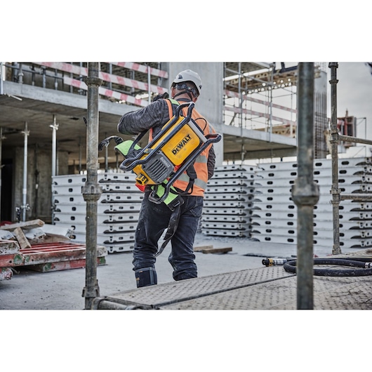 Worker carries DEWALT DCPS310N tool at construction site, wearing safety gear and orange vest.