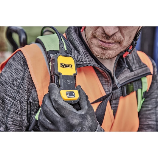 Gloved hand holding a DEWALT DCPS310N device. Person wearing a safety vest and gear outdoors.