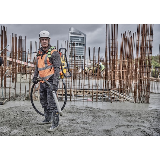 Worker uses DEWALT concrete vibrator DCPS310N at construction site, surrounded by metal rods and wet concrete.