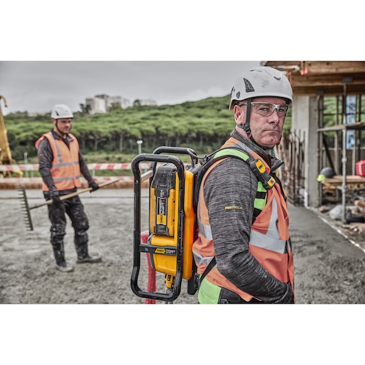 Worker in safety gear outdoors carries a yellow DEWALT tool on their back. Construction site in background.