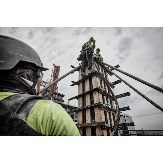 Workers in safety gear on a construction site, assembling wooden framework with pipes, under a cloudy sky.