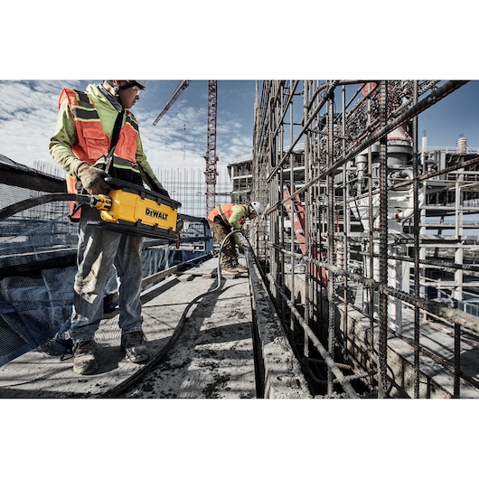 Two workers use a DEWALT tool at a construction site, surrounded by steel bars and scaffolding.