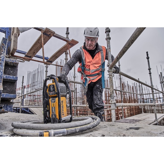 Worker in safety gear using a DEWALT cordless tool on a construction site with scaffolding and concrete.