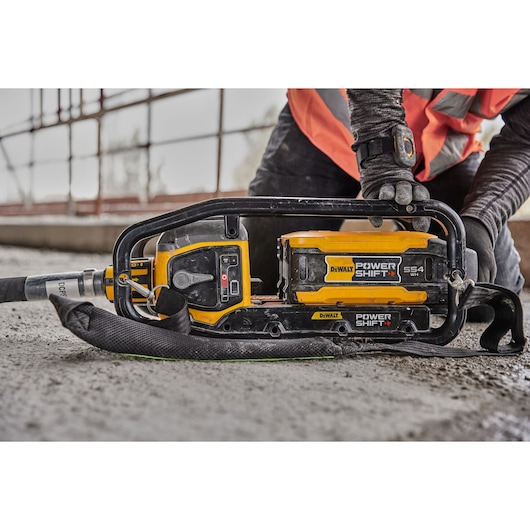 DEWALT cordless concrete vibrator tool placed on construction site ground with worker in safety gear nearby.