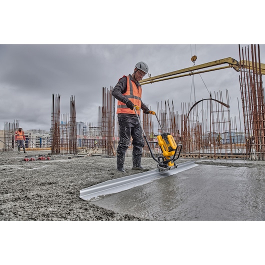 Worker uses DEWALT screed to smooth wet concrete at a construction site with steel rods and machinery visible.