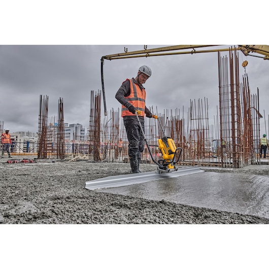 Worker uses a DEWALT power tool to smooth concrete at a construction site; steel rods are visible in the background.