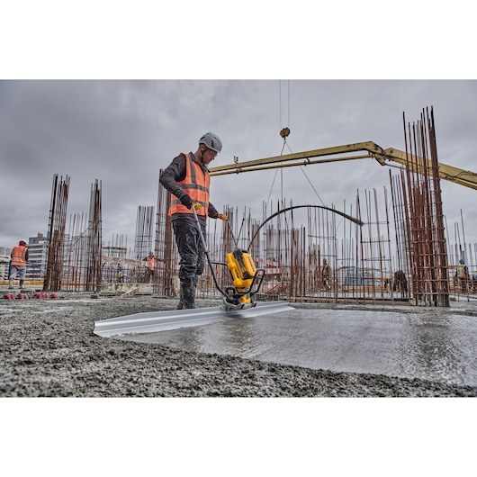 Worker uses DEWALT tool to level concrete on a construction site with steel rods and heavy machinery.