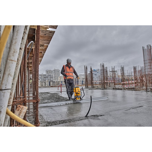 Worker using DEWALT tool to smooth concrete on construction site with buildings and steel rods in background.